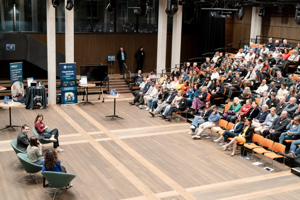 A panel of four people speaks on stage in front of a large seated audience in a modern auditorium. The audience listens attentively, and banners and tables are set up near the stage.