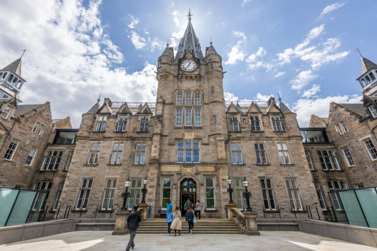 A grand, historic stone building with a central clock tower, pointed spires, and ornate windows. Several people walk up steps toward the entrance under a bright, partly cloudy sky.