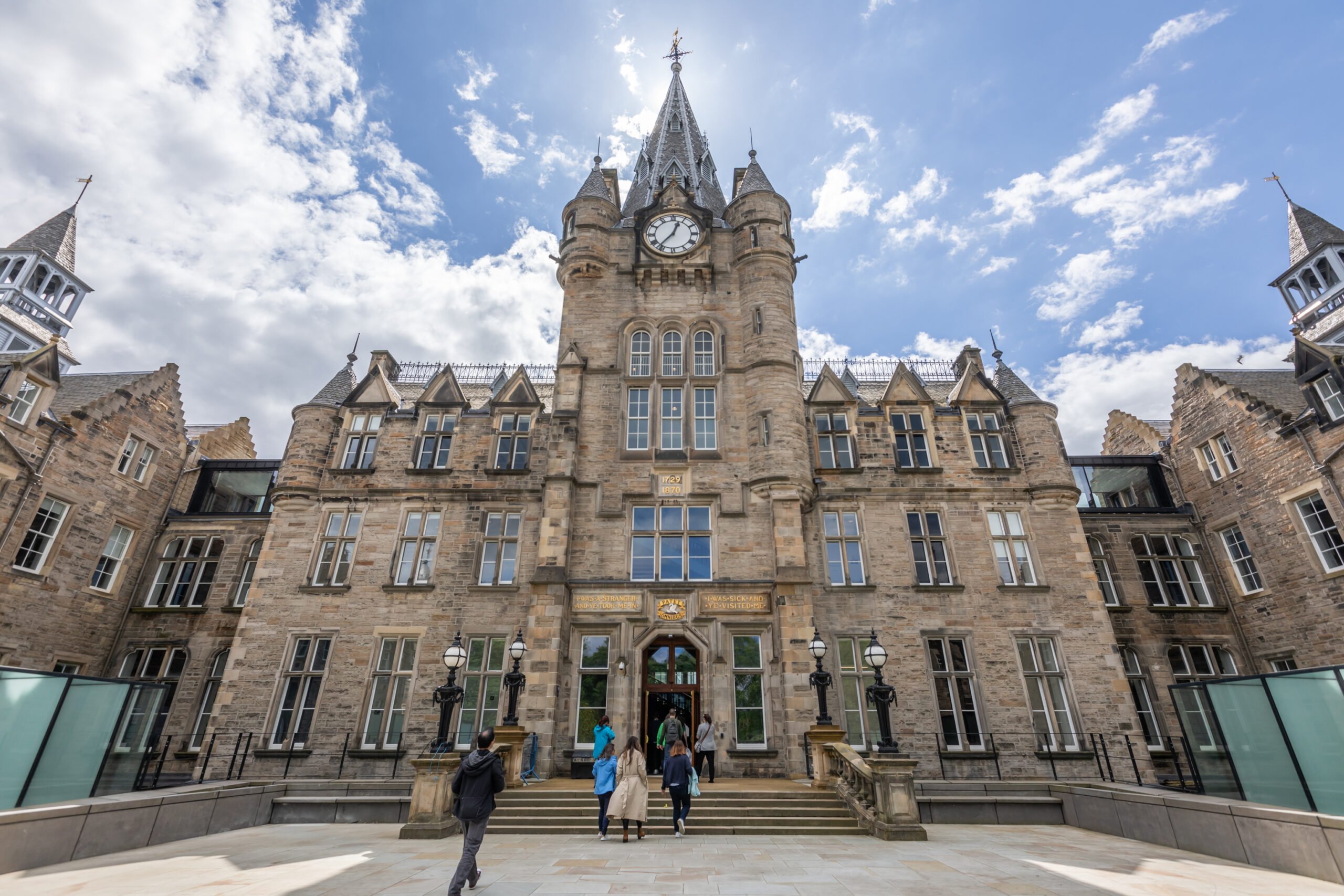 A grand, historic stone building with a central clock tower, pointed spires, and ornate windows. Several people walk up steps toward the entrance under a bright, partly cloudy sky.