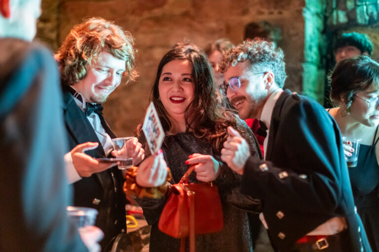 Three people dressed formally stand close together at a lively indoor event, smiling and looking at a card held by the woman in the center. Others mingle in the softly lit background.