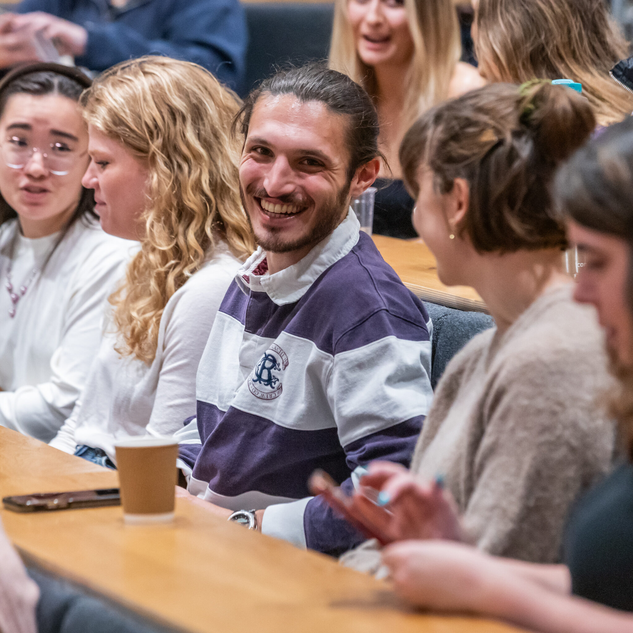 A group of people sit in a lecture hall, talking and smiling. One man in the center, wearing a striped shirt, looks directly at the camera and smiles. A paper coffee cup sits on the desk in front of them.