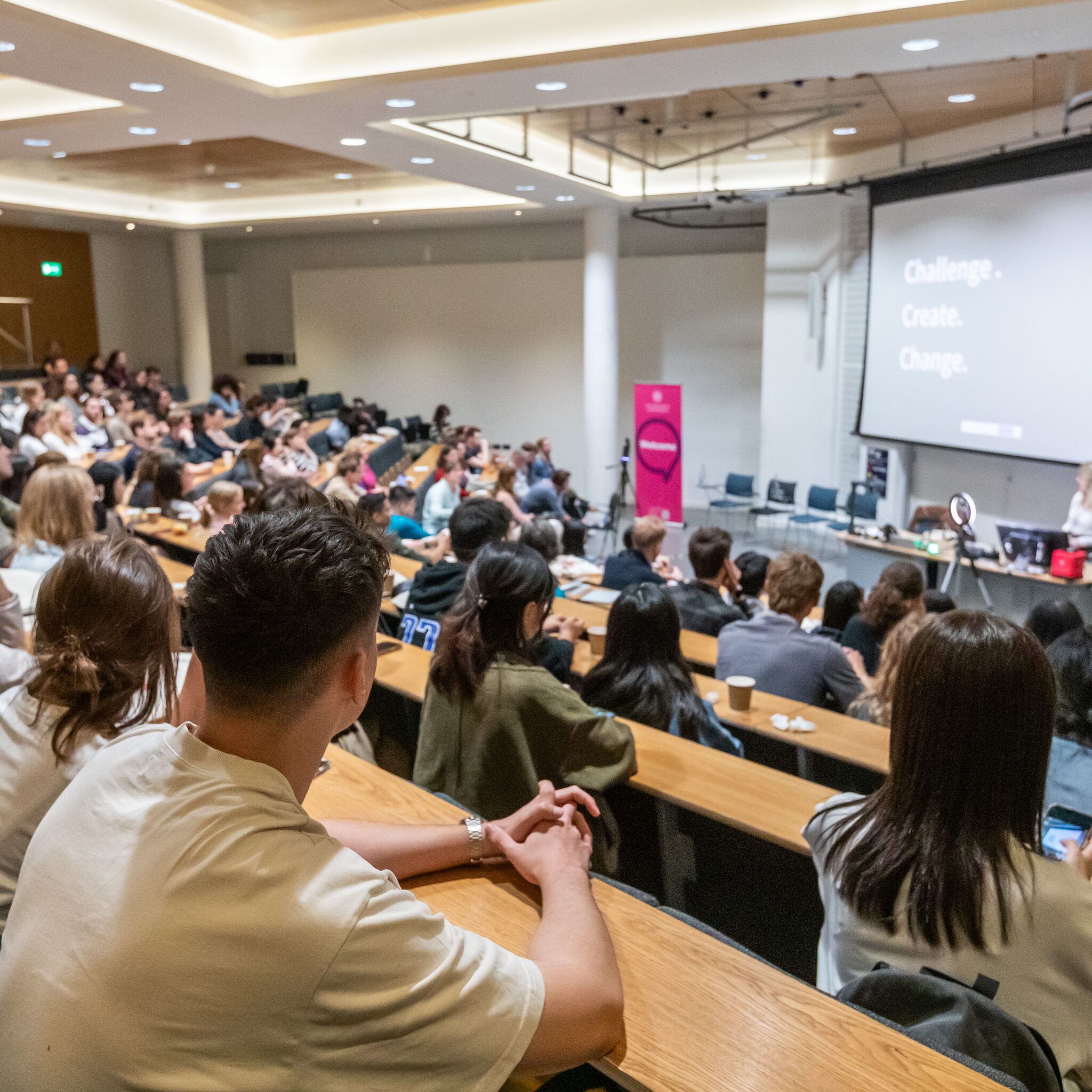 A large group of people sit in a modern lecture hall, attentively watching a speaker at the front near a screen displaying the words "Challenge. Create. Change.