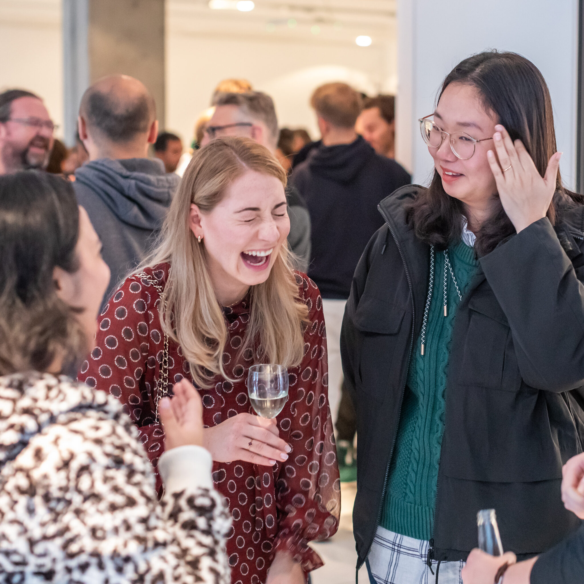 Four women stand in a lively, brightly lit room, laughing and chatting together. One holds a glass of champagne. Other people mingle in the background, creating a social, cheerful atmosphere.