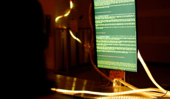 A glowing computer monitor displays green text code in a dark room, with illuminated cables or LED strips wrapped around the desk and screen. A silhouette of a person faces the monitor.