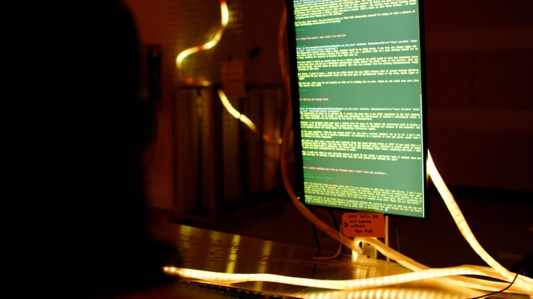 A glowing computer monitor displays green text code in a dark room, with illuminated cables or LED strips wrapped around the desk and screen. A silhouette of a person faces the monitor.