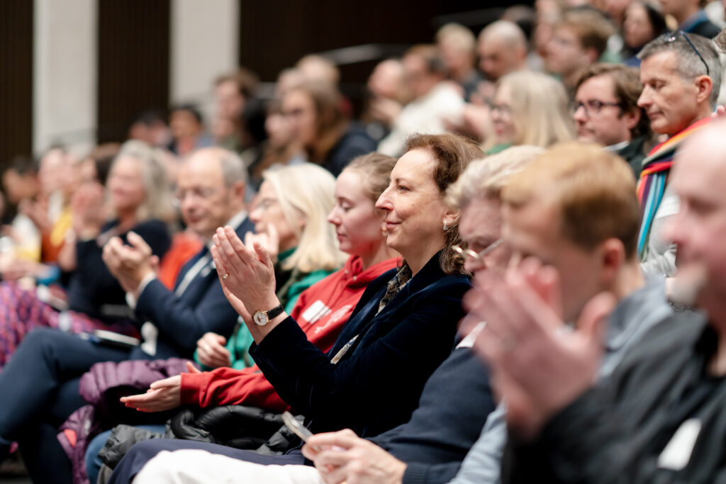 A diverse audience sits closely together in an auditorium, attentively watching and applauding an unseen speaker or performance. Most people are focused forward, some mid-clap, with expressions of interest and engagement.