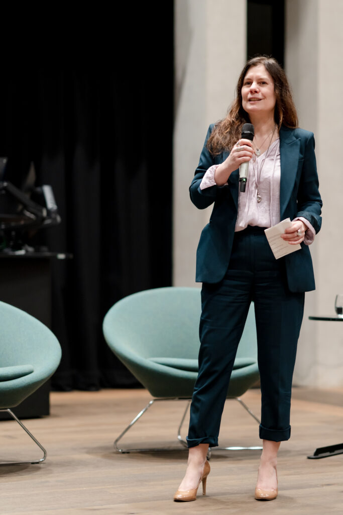A woman in a teal suit and heels stands on stage holding a microphone and note cards, speaking to an audience. Two modern green chairs and a black background are visible behind her.