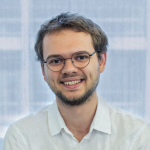 A young man with glasses, short light brown hair and a beard, wearing a white shirt, smiling in front of a blurred background with soft blue and white tones.