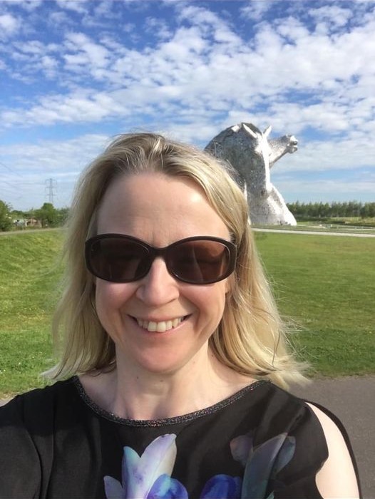 A woman with blonde hair and sunglasses smiles for a selfie outdoors, with a large silver horse-head sculpture in the background under a blue sky with scattered clouds.