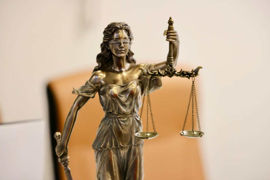 A bronze statue of Lady Justice holding balanced scales in one hand and a sword in the other, wearing a blindfold. The background is blurred and neutral in color.