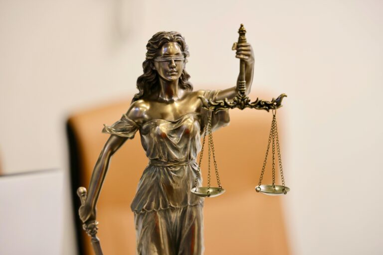 A bronze statue of Lady Justice holding balanced scales in one hand and a sword in the other, wearing a blindfold. The background is blurred and neutral in color.