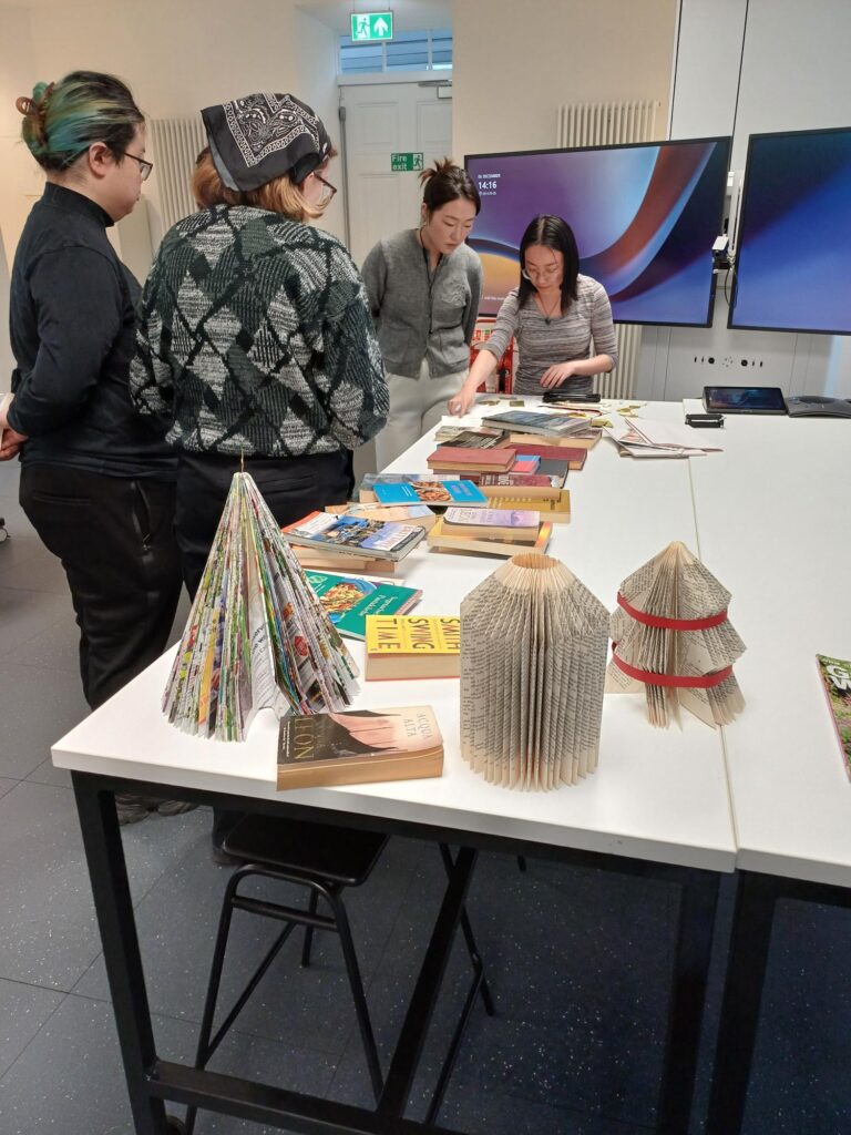 Four people stand around a table covered with books and book art, including folded paper sculptures. Two large monitors are in the background, and the setting appears to be a classroom or workshop.