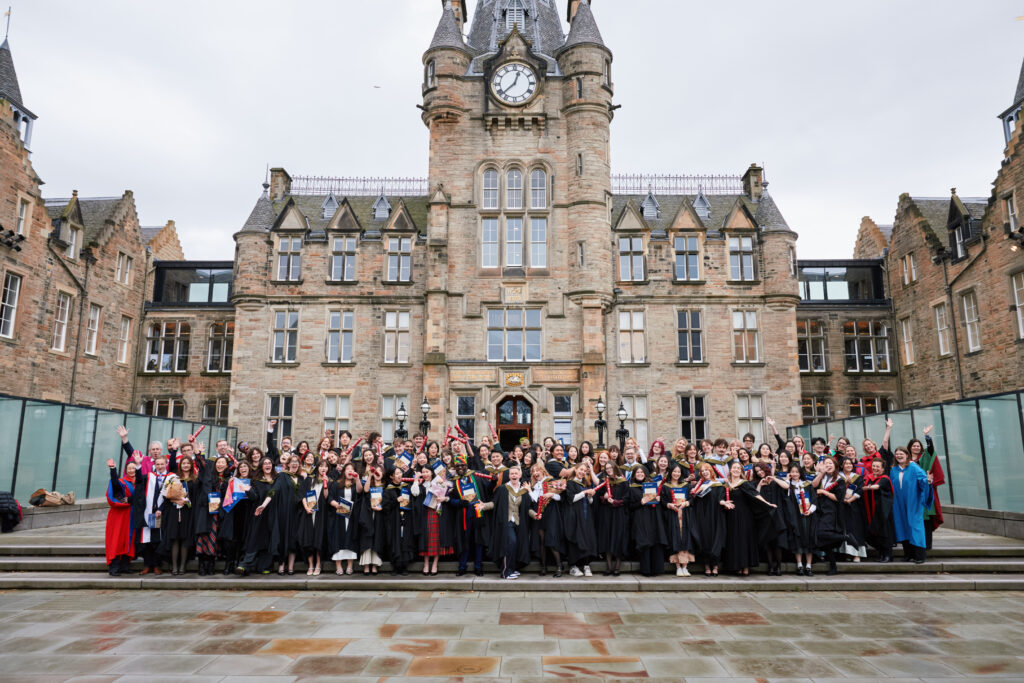 A large group of graduates in black gowns and colorful hoods pose for a photo on steps in front of a grand stone building with a clock tower on a cloudy day.