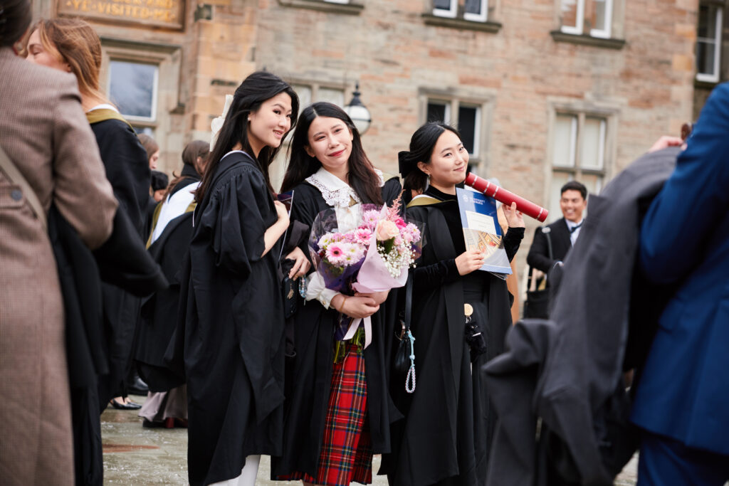 Three women in graduation gowns pose together outdoors; one holds a bouquet of flowers and another holds a diploma. Other graduates and people are visible in the background near a historic stone building.