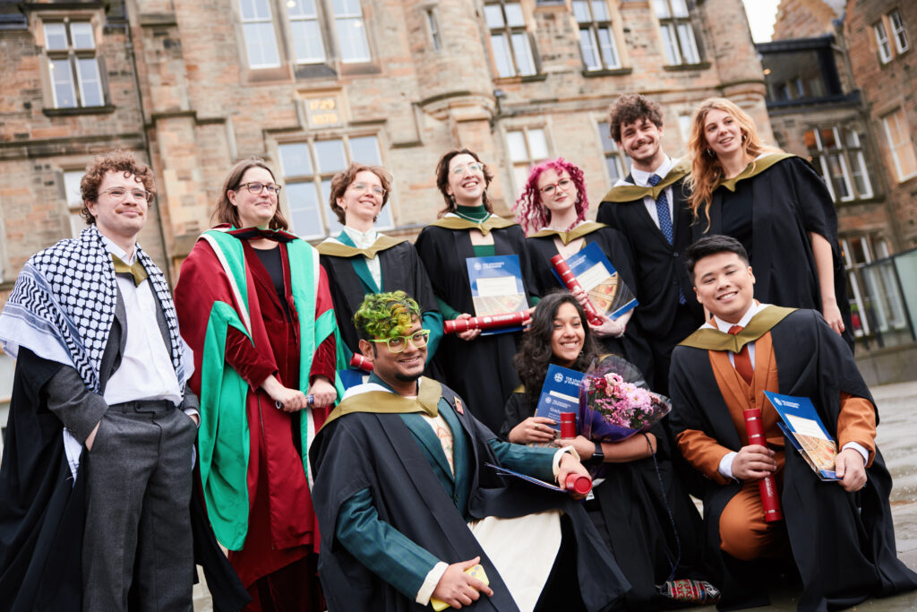 A group of graduates in academic gowns and caps pose happily outside a historic stone building, holding diplomas and flowers, smiling and celebrating their achievement.