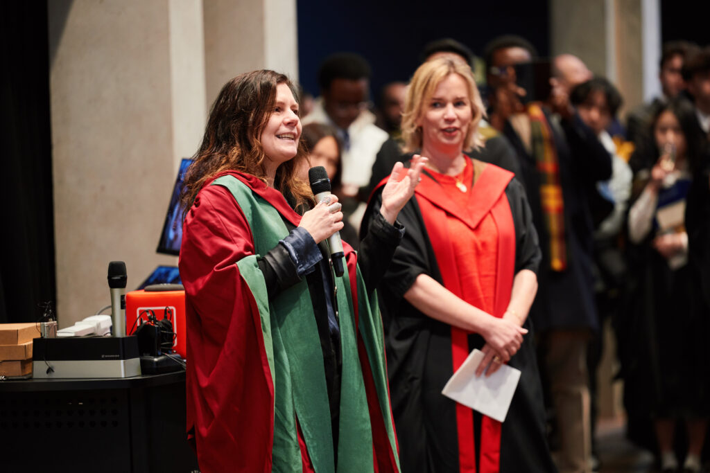 Two women in academic robes stand together; one is speaking into a microphone while the other smiles and holds papers. There is a crowd in the background at what appears to be a graduation or formal event.