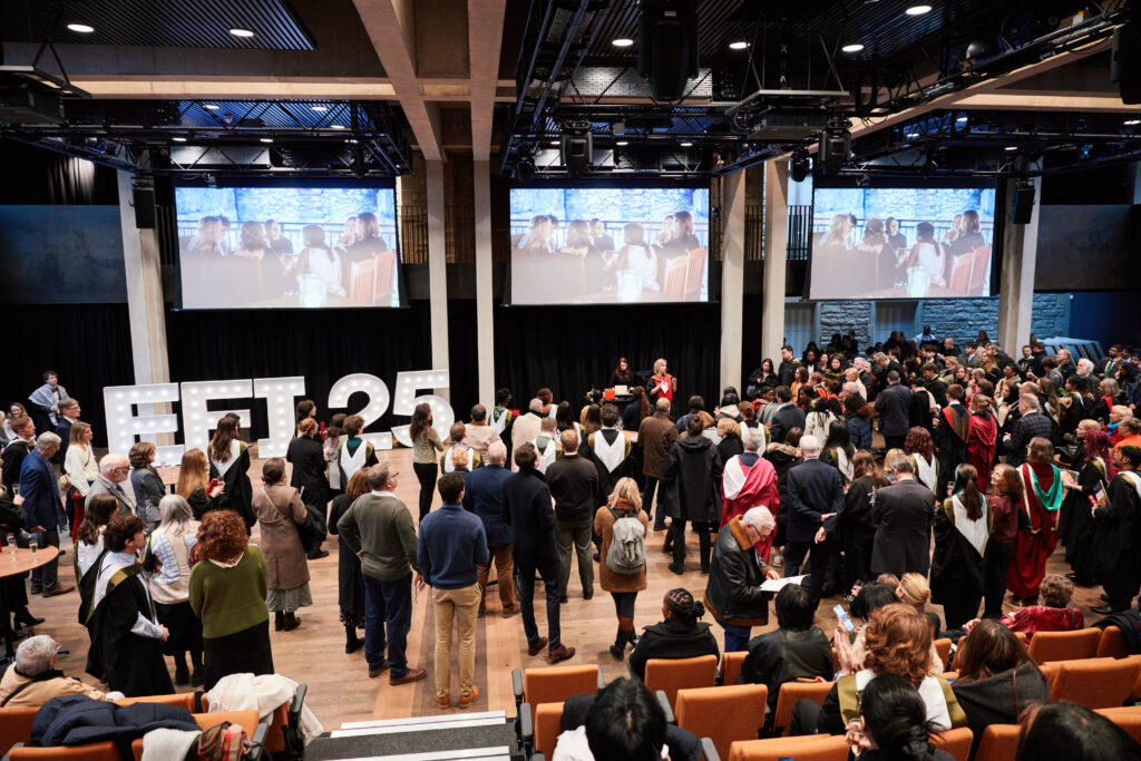 A large crowd gathers in an auditorium for a celebration, with people in academic robes and others in formal attire. White letters spelling "EFT 25" are on stage, and images are projected on large screens above.