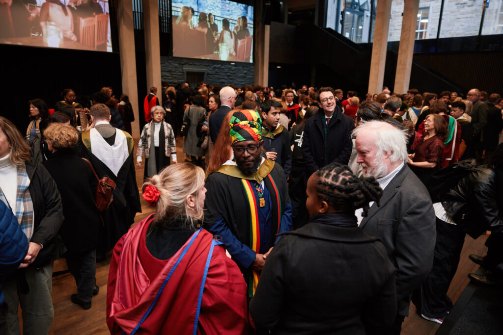 A large group of people gathers indoors at a graduation ceremony. Some are dressed in academic gowns and colorful attire, while others wear formal clothes, engaging in conversation and celebration.