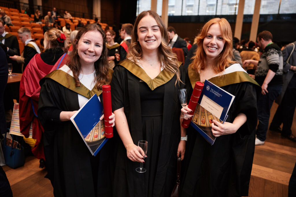 Three young women in graduation gowns and sashes smile at the camera, holding diplomas and certificates, in a crowded indoor venue with other graduates and attendees in the background.