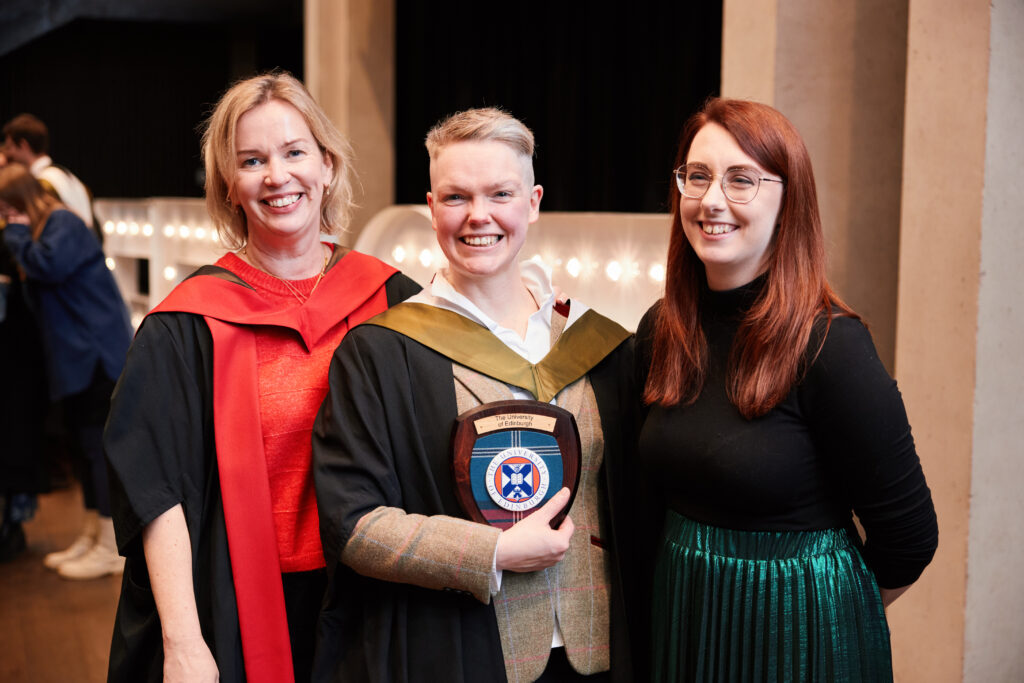 Three smiling people stand together indoors. The center person, wearing graduation robes, holds a University of Edinburgh plaque. The others stand on either side, one in academic dress and one in casual attire.