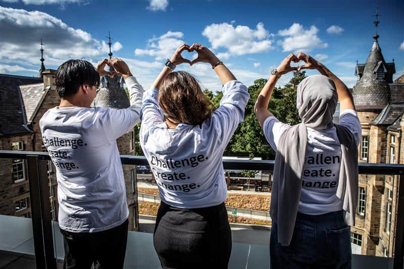 Three people stand on a balcony with their backs to the camera, wearing matching shirts that say "Challenge. Create. Change." and making heart shapes with their hands against a blue sky and historic buildings.