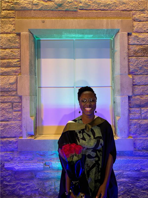 A smiling person wearing glasses and a graduation gown stands holding a bouquet of red roses in front of a stone wall and window illuminated by blue and purple lights.