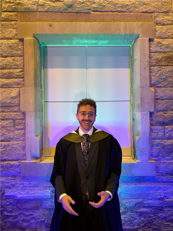 A young man wearing a graduation gown and hood stands smiling in front of a stone wall with a window, illuminated by colorful blue and purple lighting.