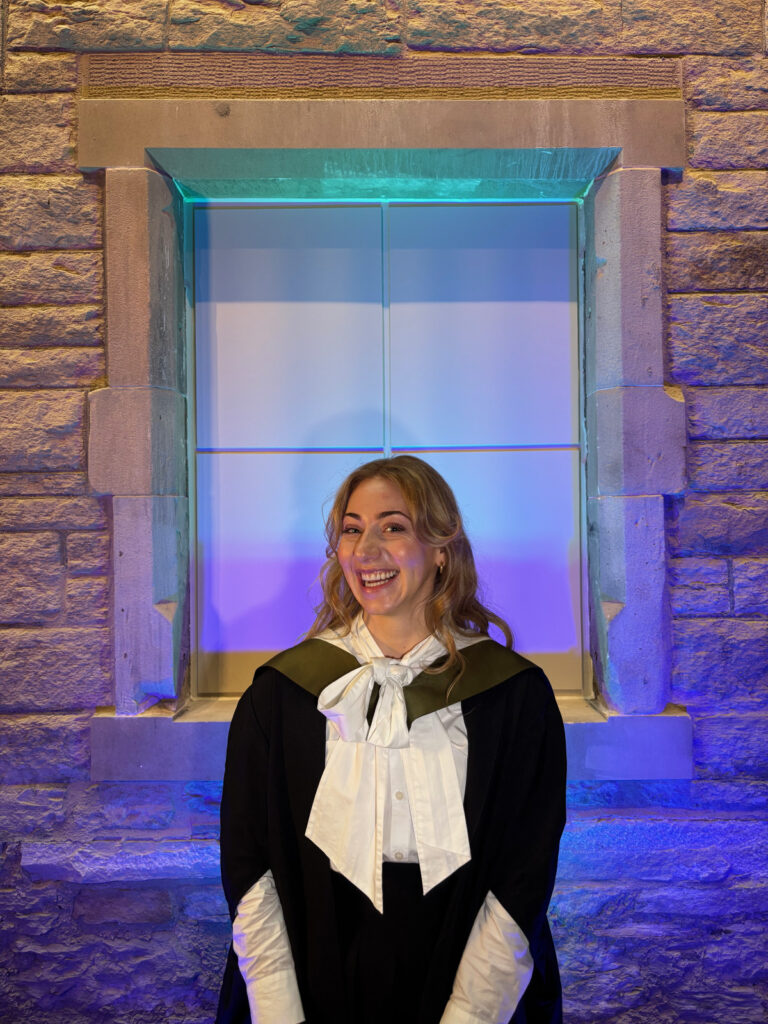 A smiling woman in academic attire stands in front of a stone wall and window, illuminated by blue and purple lights. She wears a black gown with a white shirt and a light-colored bow.