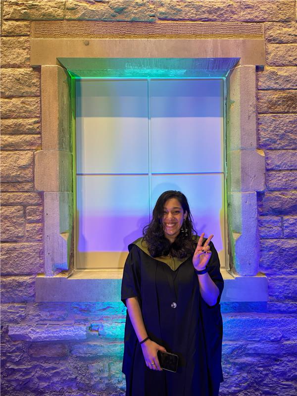 A person in a black graduation gown stands smiling in front of a stone wall and window, holding up a peace sign with one hand and a phone in the other. Colorful lights illuminate the scene.
