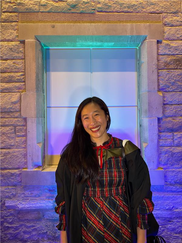 A woman with long dark hair, wearing a red and black plaid dress and a black academic gown, smiles in front of a stone wall and a brightly lit window at night.