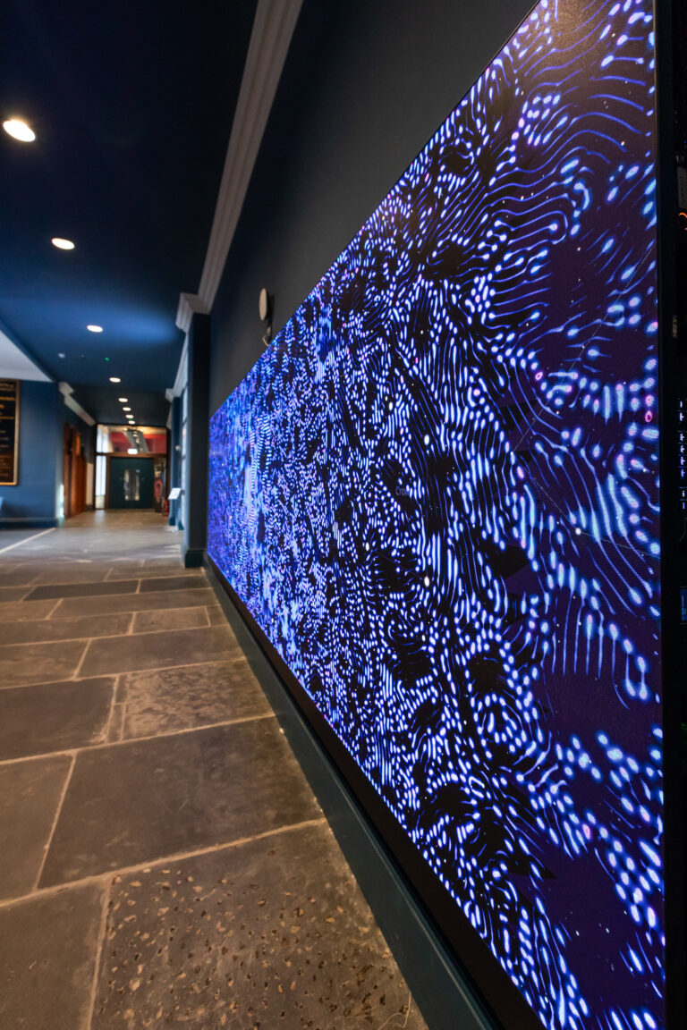 A long digital screen on a wall displays abstract blue and white wave patterns in a hallway with stone flooring and dark walls.