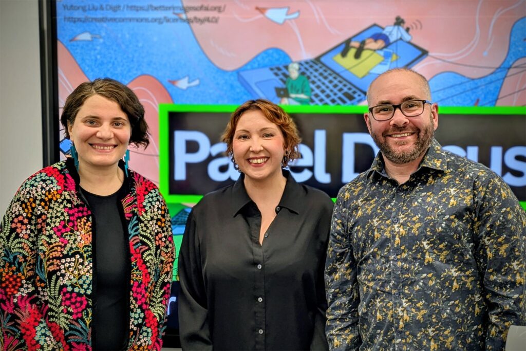 Three people stand smiling in front of a screen displaying the words “Panel Discuss.” The background shows a colorful illustration with people, digital devices, and abstract shapes.