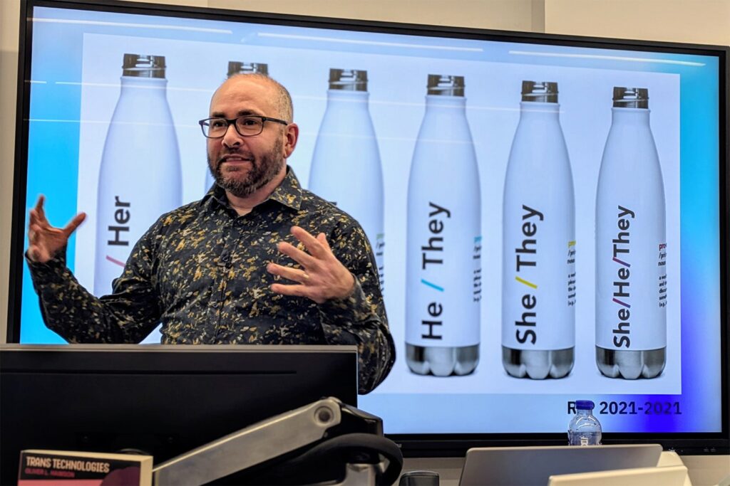 A person gestures while presenting in front of a screen displaying water bottles labeled with pronouns: "He/Her," "He/They," "She/They," and "She/He/They." The year "2021-2021" appears on the slide.