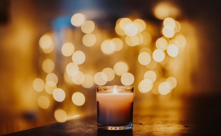 A lit candle in a glass holder sits on a surface, with warm, golden bokeh lights softly blurred in the background, creating a cozy and serene atmosphere.