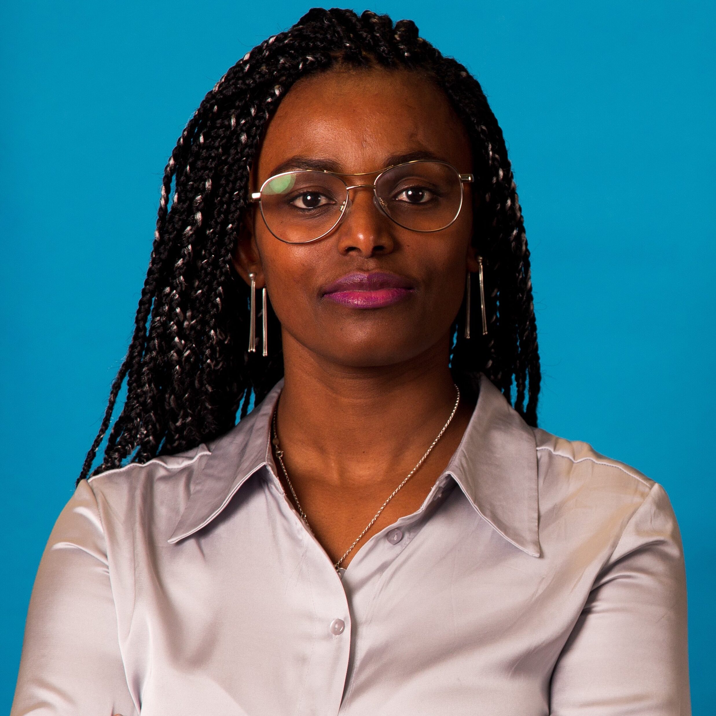 A woman with braided hair, wearing glasses, a silver satin blouse, and earrings, stands against a bright blue background, looking confidently at the camera with her arms crossed.