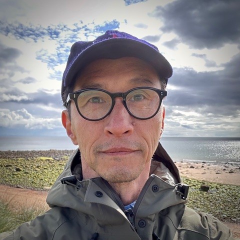 A person wearing glasses, a dark cap, and a green jacket stands outdoors by a rocky shoreline with the sea and a cloudy sky in the background.