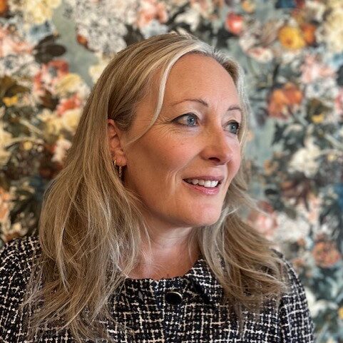 A woman with long blonde hair and a black-and-white patterned jacket smiles, looking to the right. Behind her is a colorful, floral-patterned background with various flowers and leaves.