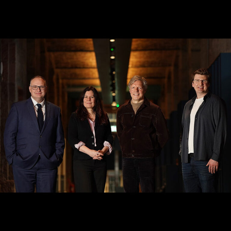 Four adults stand side by side indoors, posing and smiling at the camera. The group includes three men and one woman, all dressed in business or smart casual attire, with a softly lit hallway in the background.