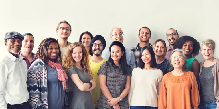 A diverse group of people of various ages and backgrounds stand together in front of a plain white wall, smiling and posing for a group photo.