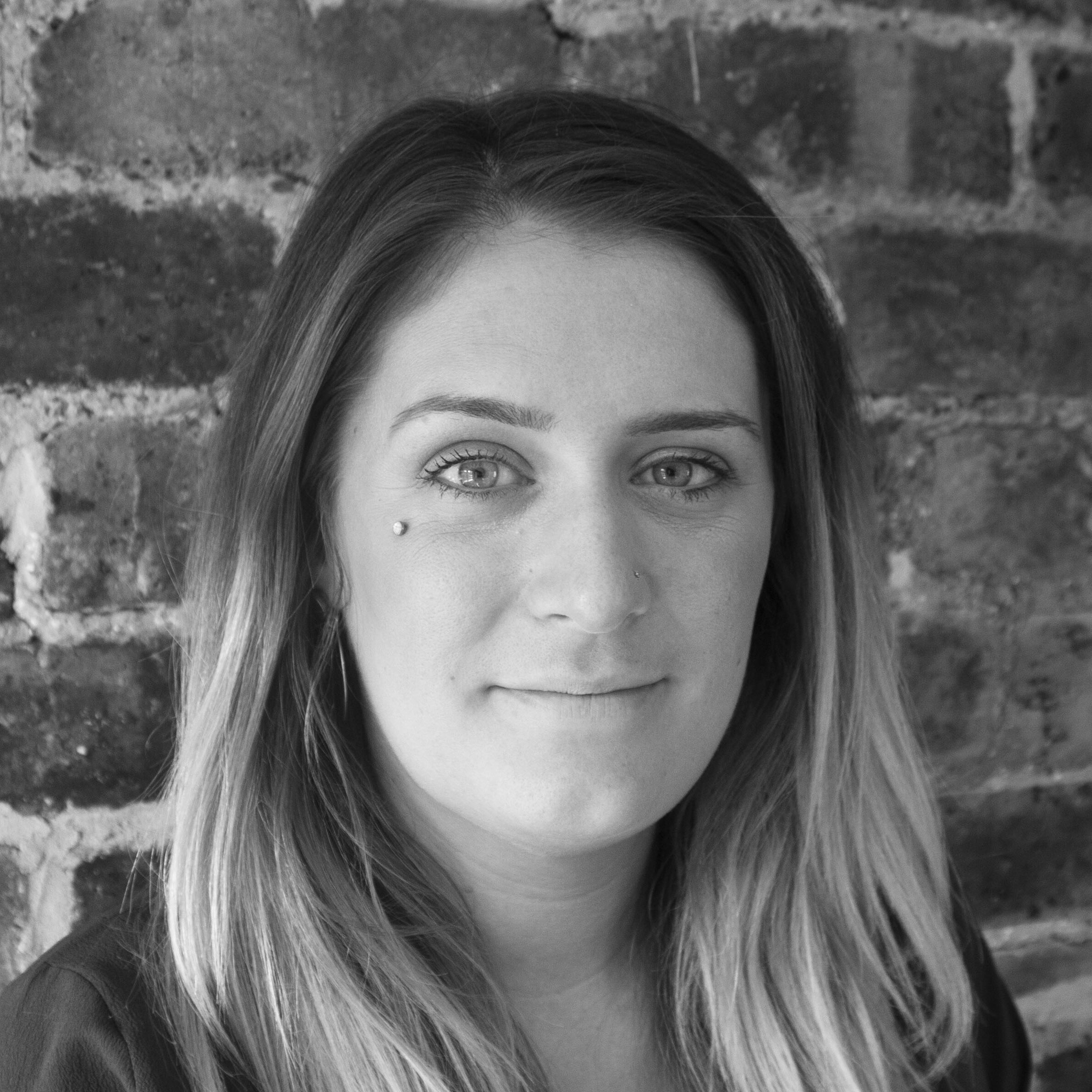 A woman with long, straight hair and a nose ring smiles slightly at the camera. She has a facial piercing on her cheek and is standing in front of a brick wall. The photo is in black and white.