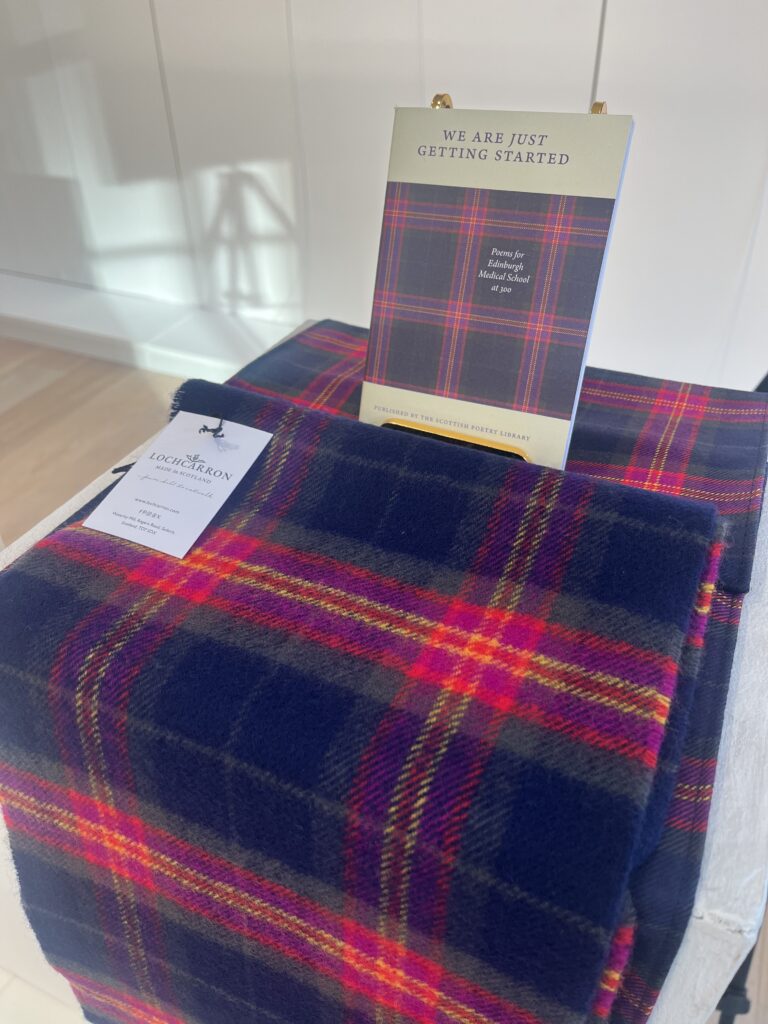 A dark blue and red plaid fabric is draped over a white surface. On top, there is a business card for "Lochcarron" and a booklet titled "We Are Just Getting Started" featuring a matching plaid design.