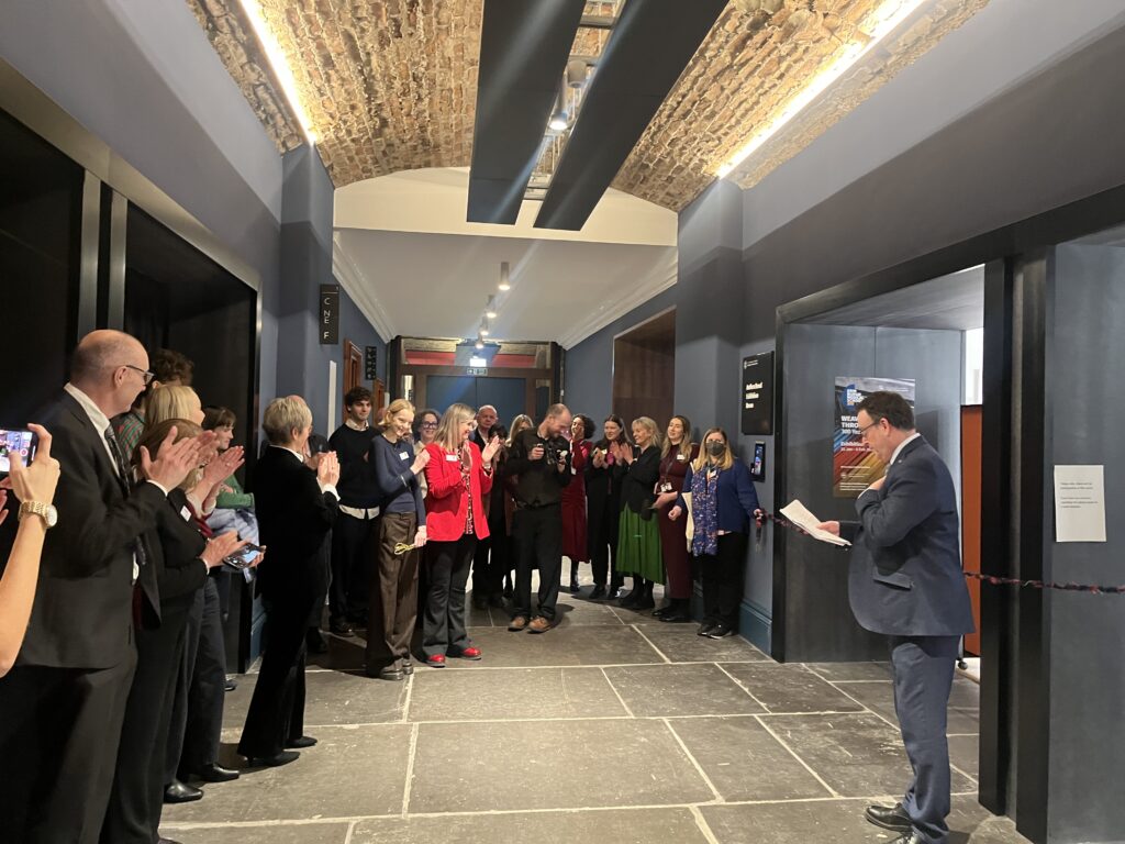 A group of people stands in a hallway with exposed brick ceiling, applauding a man in a suit who is speaking and holding papers. Most are dressed in business attire and some are taking photos. The atmosphere appears celebratory.