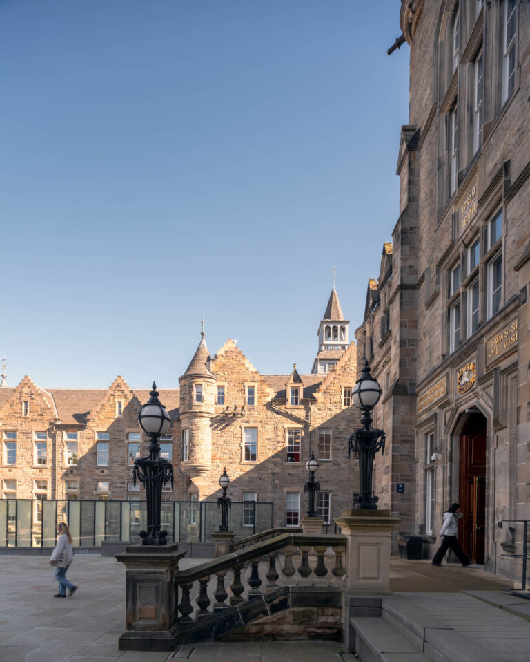 A historic stone university building with arched windows and a staircase, under a clear blue sky. Two people are walking in the courtyard near ornate lamp-posts; the building features turrets and a clock tower in the background.