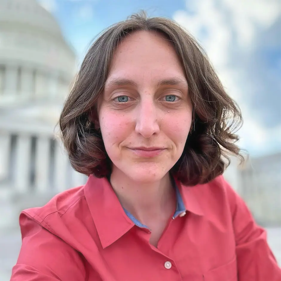 A person with wavy brown hair and blue eyes, wearing a red button-up shirt, takes a selfie outdoors in front of a white domed building under a partly cloudy sky.
