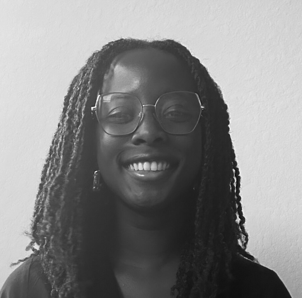 Black and white photo of a person with long, twisted hair wearing glasses and smiling, against a plain, light-coloured background.