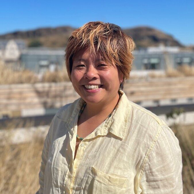 A person with short, light brown hair and a light-coloured button-up shirt smiles outdoors, with dry grass and buildings visible in the background under a clear blue sky.