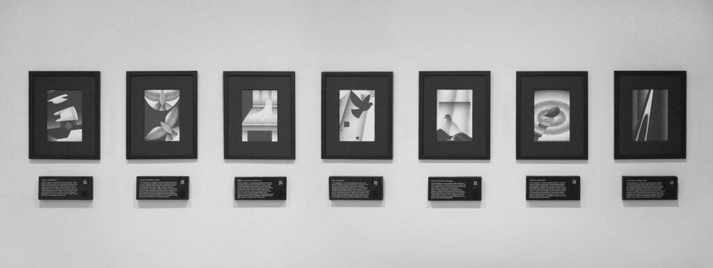 Seven framed abstract black and white artworks hang in a row on a gallery wall, each with a descriptive plaque beneath it. The images feature geometric and organic shapes.