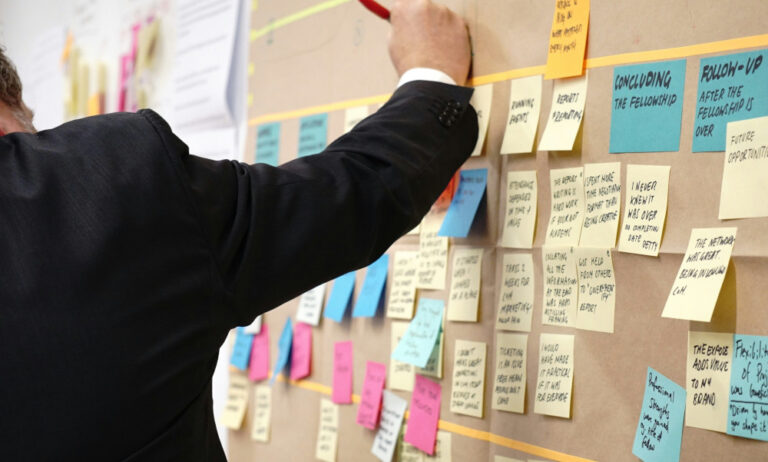 A person in a suit writes on a large board covered with colourful sticky notes and papers, each containing handwritten notes, tasks, and ideas, suggesting a brainstorming or project planning session.