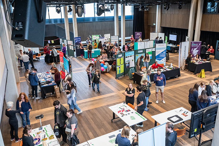 Large indoor hall with stands and tables set up for a careers or information fair. People are walking around, talking to exhibitors, and gathering materials. The space is bright with natural light from tall windows.