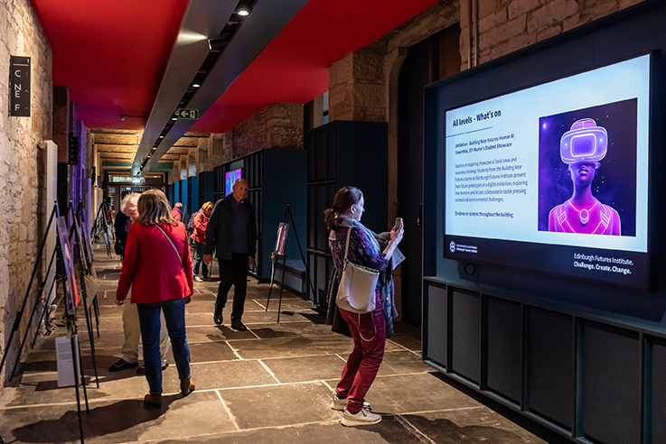 People view interactive digital displays in a modern exhibition corridor with stone walls and a red ceiling. One woman takes a photo of a screen featuring a person wearing a virtual reality headset.
